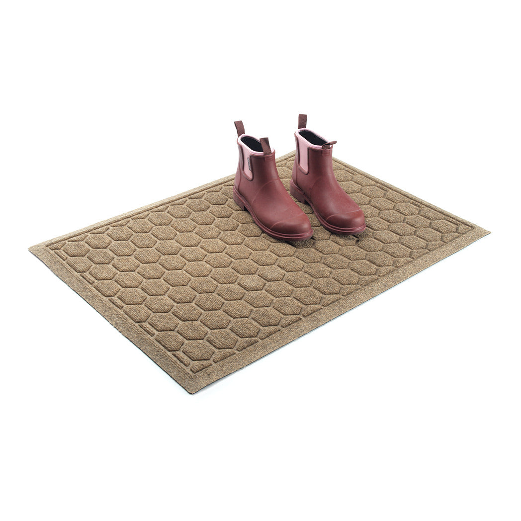 A WaterHog Honeycomb outdoor doormat in tan, featuring a hexagonal pattern displayed on a white background, with boots placed on top of the mat.