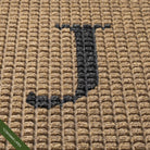 Close-up of the hand-cut and inlaid serif 'J' in charcoal on a tan WaterHog squares doormat; latex disclaimer