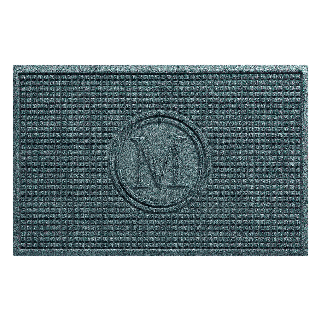 Overhead of traditional Squares WaterHog indoor/outdoor doormat monogrammed with the letter M, in a light blue/grey color, on a stain-resistant surface.