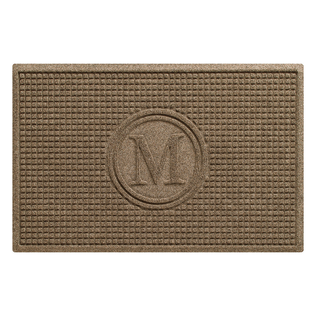 An overhead of a WaterHog Squares doormat monogrammed with an M in the center with a circle around it, shown in a wheaty yellow and uniform grid bi-level design.