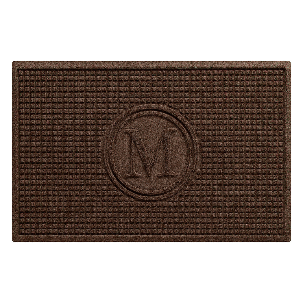 An above shot of a WaterHog Squares modern doormat, monogrammed with the letter M encased in a circle, surrounded by a uniform square grid design, shown in a dark, deep grey.