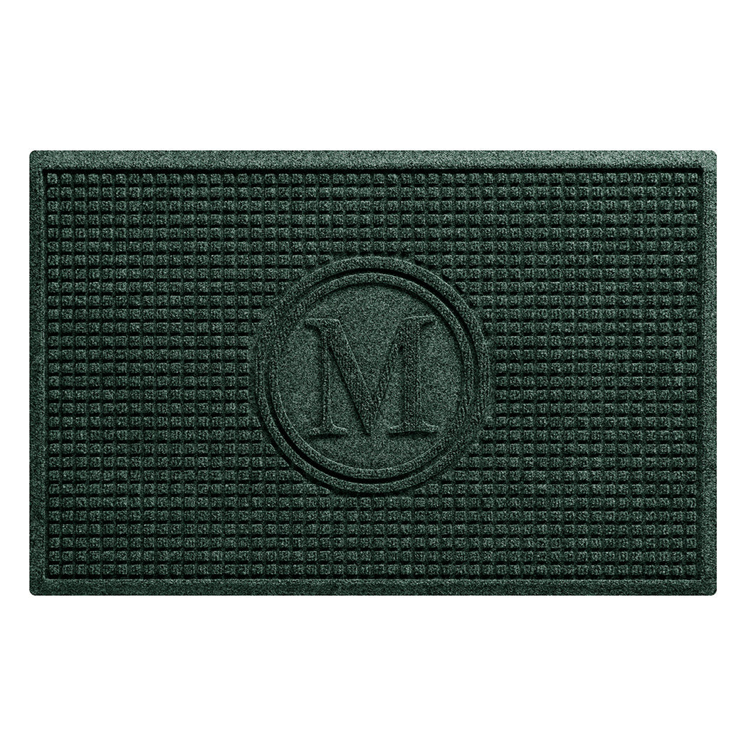 An overhead of a WaterHog Squares doormat monogrammed with an M in the center with a circle around it, shown in a deep green durable surface.