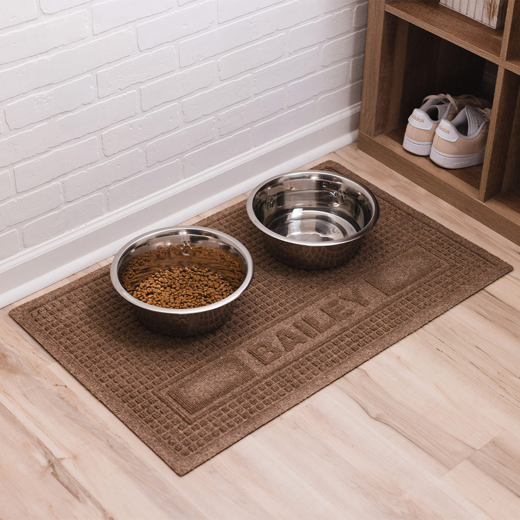 A personalized Squares pet mat with the name Bailey with a water and food dish on top located in a mud room.