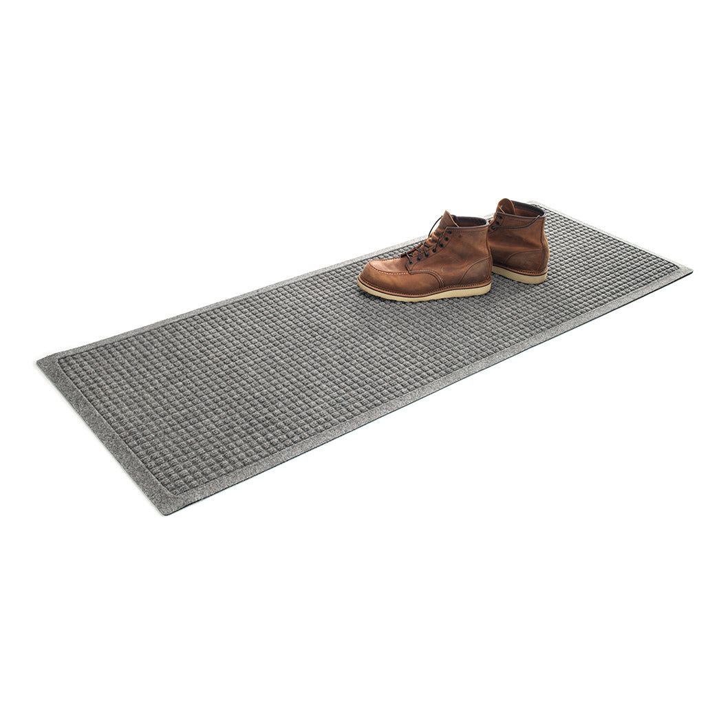 Brown boots placed on top of a light grey WaterHog runner mat with classic repeating squares pattern in a recycled PET surface and rubber backing. 