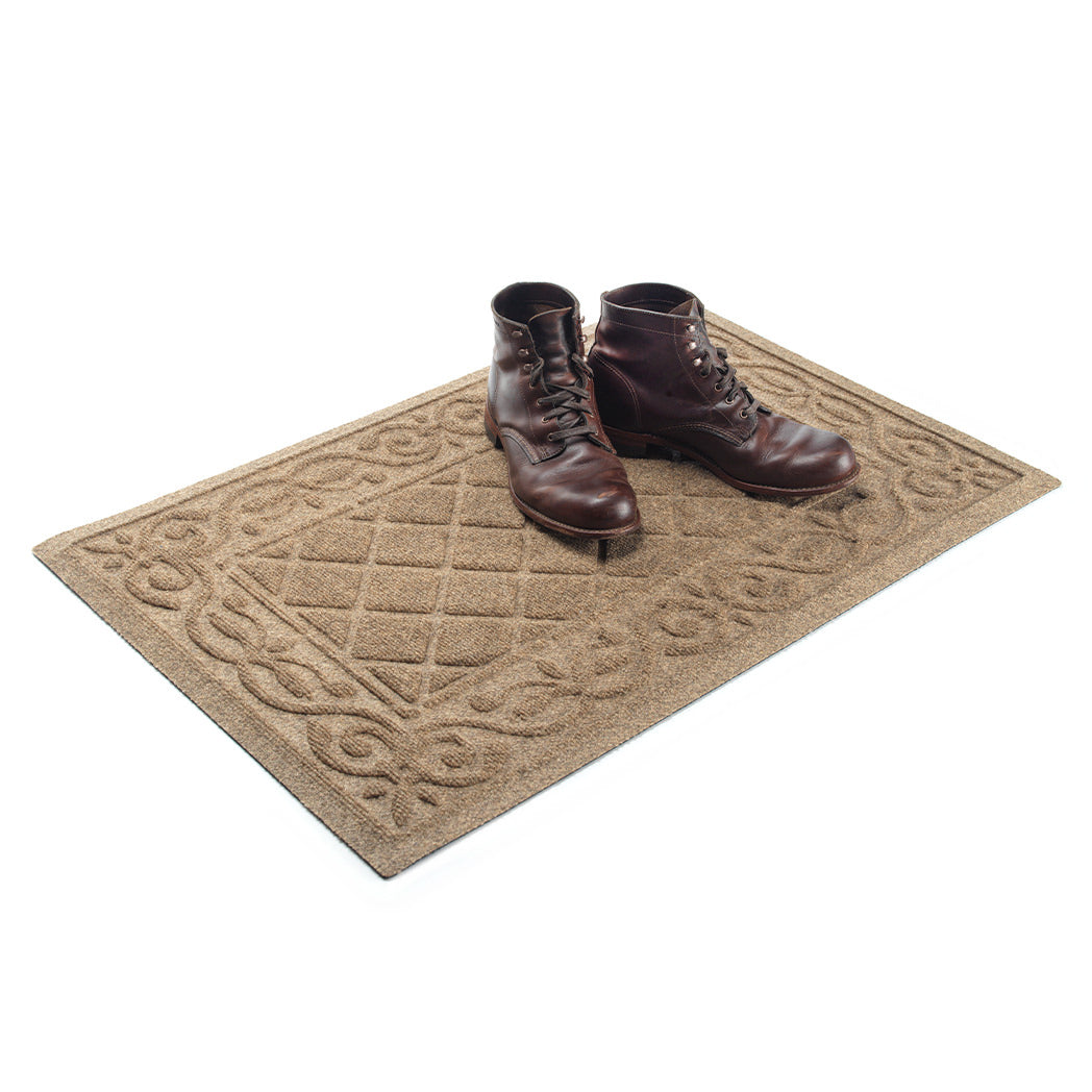 A pair of men's brown leather boots sitting on the right half of a 2'x3' WaterHog Tristan doormat in the color tan, with the other side open to fit one or two more pairs of shoes.